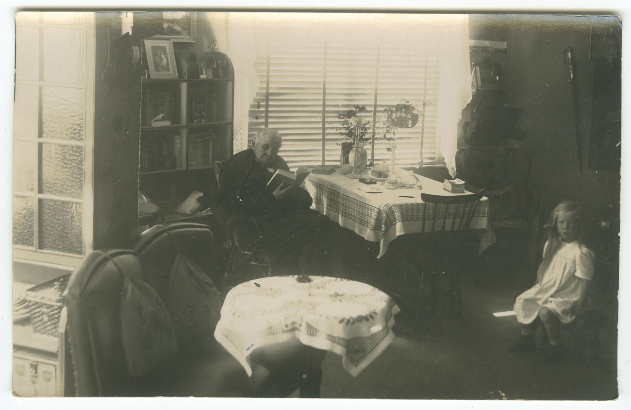svartvitt foto. Kvinna i 70 års åldern sitter på en mörk stol bredvid ett matbord med duk och läser en bok. På en pall snett till höger sitter en flicka i fem års åldern. 
