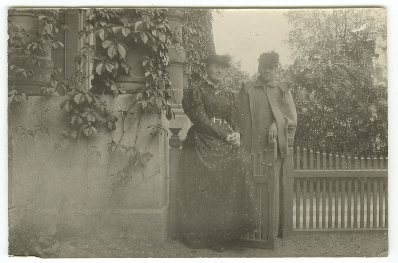 Svartvitt foto. Två kvinnor står utomhus i vid en grind till ett staket. Kvinnorna har kortklippt hår och bär mörka hattar från tidigt 1900-tal.
