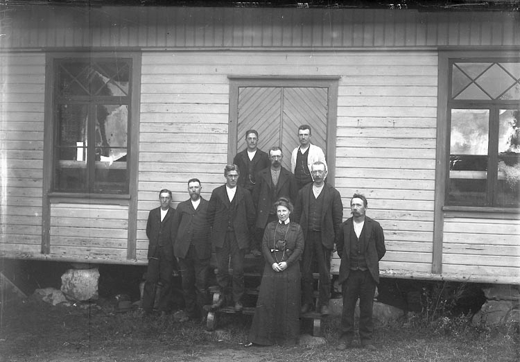 Svartvitt foto. 9 personer står på och bredvid en trappa framför ett vitt trähus byggt under tidigt 1800-tal. åtta män bär kostym och står runt en kvinna i mörk blus och kjol.