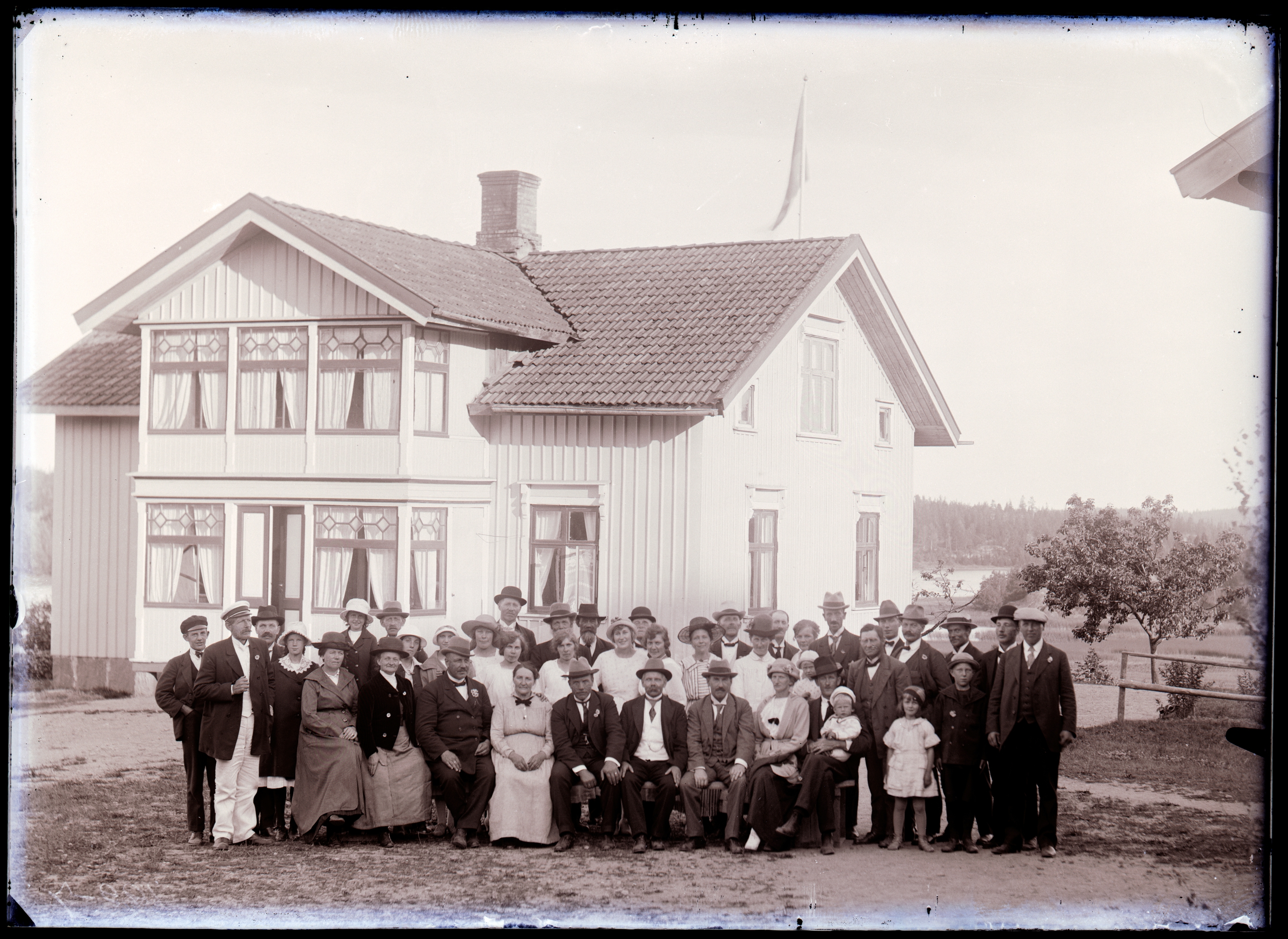 svartvitt foto. Gruppbild med 41 personer stående framför ett vitt sekelskiftshus med spröjsade flaggfönster.